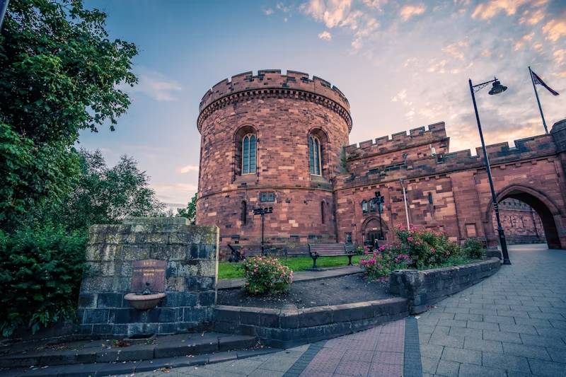 Historic stone castle tower surrounded by greenery and flowers under a colourful sky, with pathways and benches nearby.