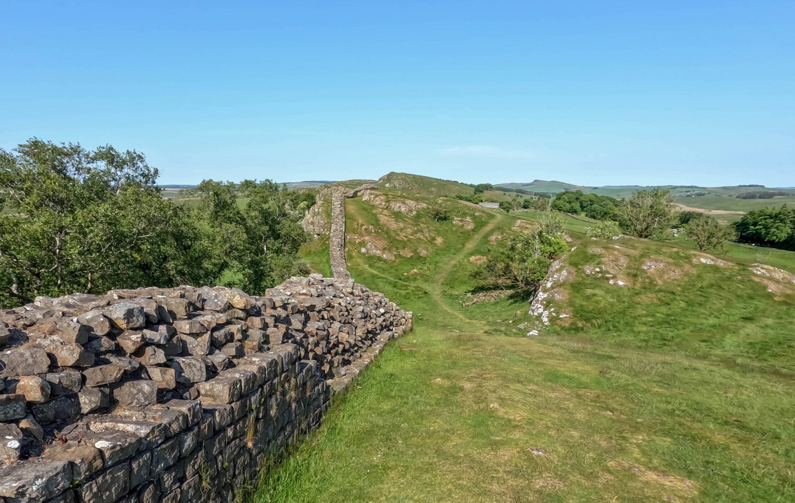 ray-harrington-anPeDd4OmeM-unsplash (1) Stone walls of an ancient structure wind through lush green hills beneath a clear blue sky, surrounded by trees and rolling landscapes.